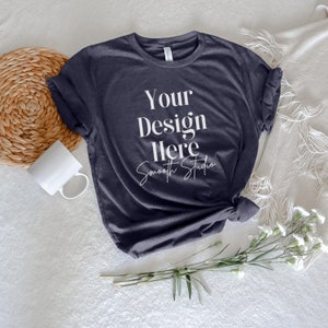 May include: A dark blue t-shirt with the text "Your Design Here" and "Smooth Studio" written in white. The t-shirt is laid flat on a white surface with a woven basket, a white mug, and a bouquet of white flowers.