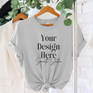 May include: A light gray t-shirt with short sleeves hanging on a wooden hanger. The shirt has the text "Your Design Here" and "Smooth Studio" written on it.