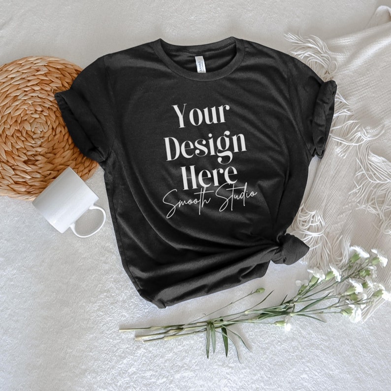 May include: A black t-shirt with the text "Your Design Here Smooth Studio" printed on the front. The t-shirt is lying on a white surface with a woven placemat, a white mug, and a bouquet of white flowers.