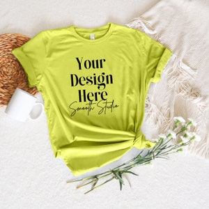 Op de afbeelding: Een felgeel T-shirt met de tekst "Your Design Here Smooth Studio" bedrukt op de voorkant. Het shirt ligt plat op een wit oppervlak met een wit deken, een rieten mand, een witte mok en witte bloemen.