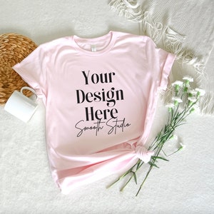 Puede incluir: Una camiseta rosa con el texto "Your Design Here" y "Smooth Studio" escrito en cursiva. La camiseta está colocada sobre una superficie blanca con una cesta de mimbre y una taza blanca a la izquierda y un ramo de flores blancas a la derecha.