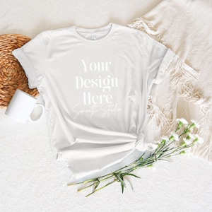 May include: A white t-shirt with the text "Your Design Here" and "Smooth Studio" written in white on the front. The t-shirt is lying on a white surface with a wicker basket, a white mug, and a bouquet of white flowers.