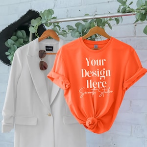 Puede incluir: Una camiseta naranja brillante con el texto "Your Design Here Smooth Studio" impreso en la parte delantera. La camiseta está colgada de una percha de madera.