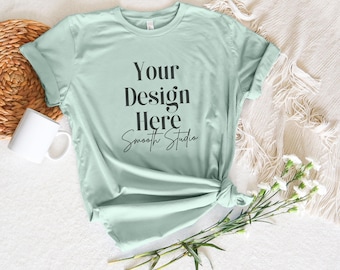 May include: A light green t-shirt with the text "Your Design Here Smooth Studio" printed on the front. The t-shirt is laid flat on a white surface with a wicker basket, a white mug, a white blanket, and white flowers.