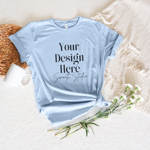 May include: A light blue t-shirt with the text "Your Design Here Smooth Studio" printed on the front. The shirt is laid flat on a white surface with a wicker basket, a white mug, a white blanket, and white flowers.