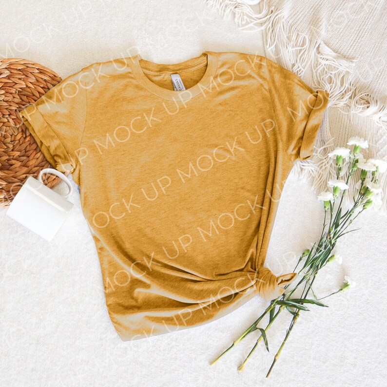 May include: A mustard yellow t-shirt with a relaxed fit, lying on a white surface with a woven basket and white flowers.