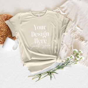 May include: A beige t-shirt with the text "Your Design Here" and "Smooth Studio" printed on the front. The t-shirt is laid flat on a white surface with a wicker basket, a white mug, and a bouquet of white flowers.
