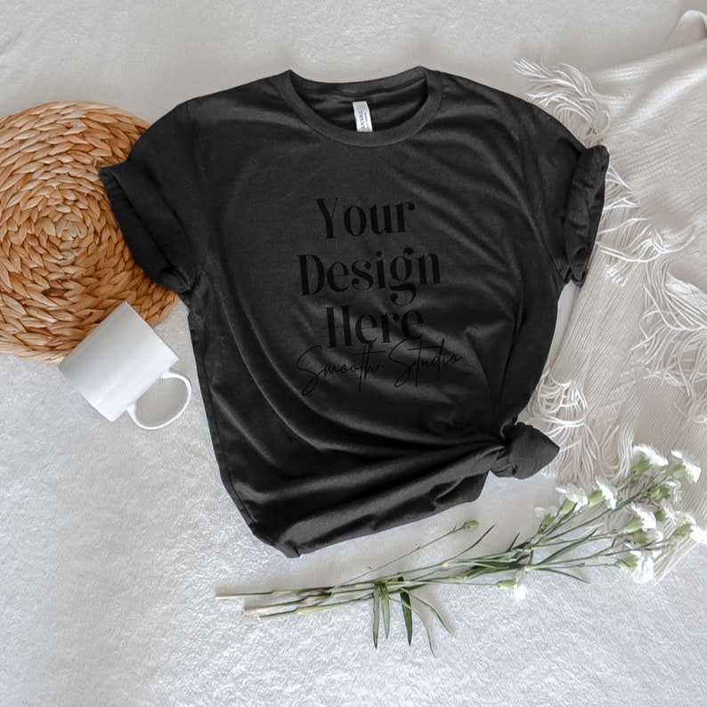 May include: A black t-shirt with the text "Your Design Here" and "Smooth Studio" printed on the front. The t-shirt is lying flat on a white surface with a wicker coaster, a white mug, and a bouquet of white flowers.