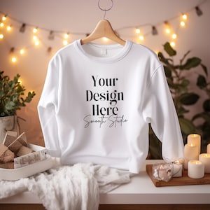 May include: A white crewneck sweatshirt hangs on a wooden hanger. The sweatshirt features the text "Your Design Here Smooth Studio" in black. The background includes soft lighting and decorative elements.