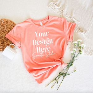 May include: A coral pink t-shirt with a white design that says "Your Design Here Smooth Studio" on a white background. The t-shirt is lying on a white surface with a wicker basket, a white mug, a white blanket, and white flowers.