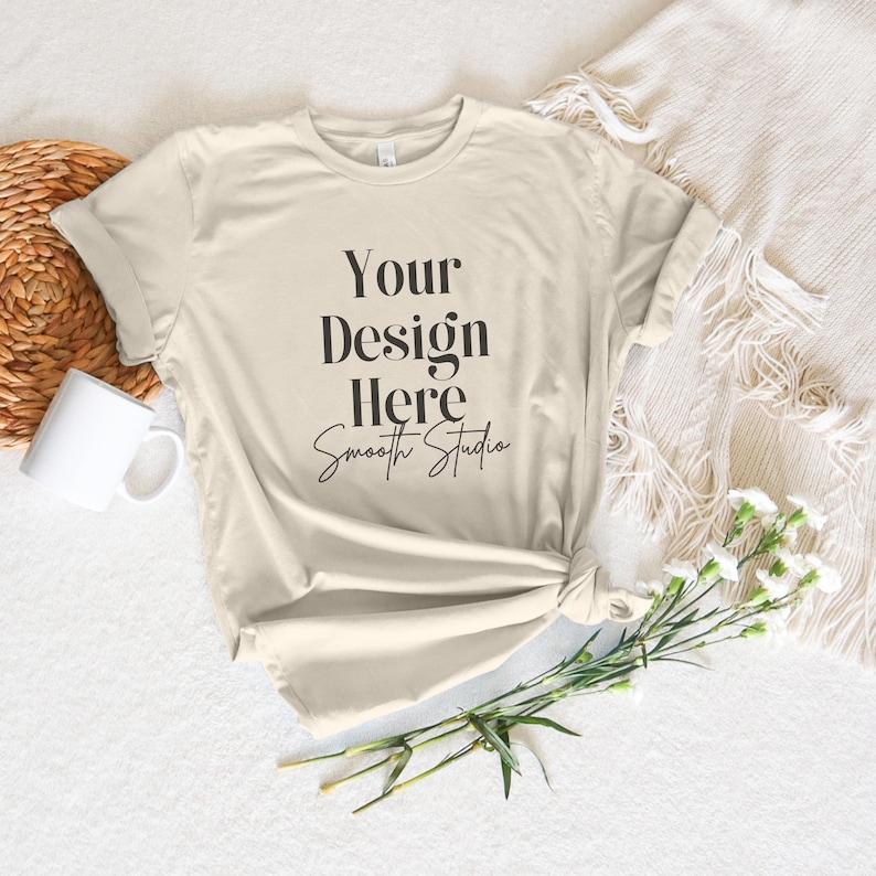 May include: A beige t-shirt with the text "Your Design Here Smooth Studio" printed on the front. The shirt is laid flat on a white surface with a wicker basket, a white mug, and a bouquet of white flowers.