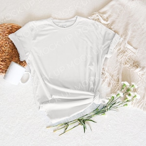 May include: A white t-shirt with rolled up sleeves is laid out on a white surface with a woven basket, a white mug, and a bouquet of white flowers.