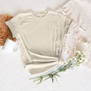 May include: A light beige t-shirt with rolled up sleeves, lying on a white surface with a woven basket, a white mug, and a bouquet of white flowers.