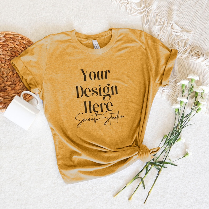 May include: A mustard yellow t-shirt with the text "Your Design Here Smooth Studio" printed on the front. The t-shirt is lying on a white surface with a wicker basket and a white mug.