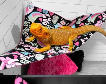 May include: A bright orange bearded dragon lizard resting in a black and pink hammock with a colorful print of hearts, stars, and musical notes. The hammock is hanging from a white tiled wall.