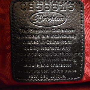 K&ouml;nnte beinhalten: Nahaufnahme eines schwarzen Lederetiketts mit dem Brighton-Logo und dem Text "The Brighton Collection". Das Etikett hat oben die Nummer "C856615". Der Hintergrund ist rot.