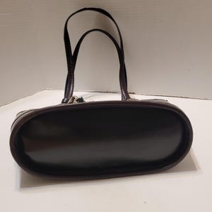K&ouml;nnte beinhalten: Eine dunkelbraune Handtasche mit gl&auml;nzendem Finish. Die Tasche hat zwei gew&ouml;lbte Griffe und eine abgerundete rechteckige Form. Die Griffe sind in einem dunkleren Braunton gehalten und scheinen strukturiert zu sein. Die Tasche steht auf einer wei&szlig;en Oberfl&auml;che.