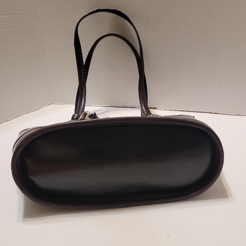 K&ouml;nnte beinhalten: Eine dunkelbraune Handtasche mit gl&auml;nzendem Finish. Die Tasche hat zwei gew&ouml;lbte Griffe und eine abgerundete rechteckige Form. Die Griffe sind in einem dunkleren Braunton gehalten und scheinen strukturiert zu sein. Die Tasche steht auf einer wei&szlig;en Oberfl&auml;che.