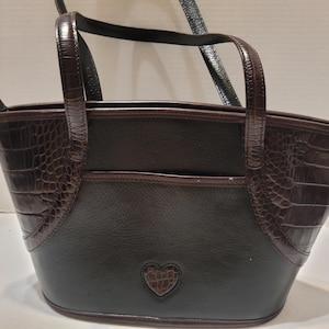 K&ouml;nnte beinhalten: Eine braune und schwarze Handtasche mit herzf&ouml;rmigem Detail. Die Tasche hat ein strukturiertes Krokodilmuster an den Seiten und Griffen, mit einem glatten, dunkelbraunen K&ouml;rper. Sie hat zwei Griffe und einen Schultergurt.