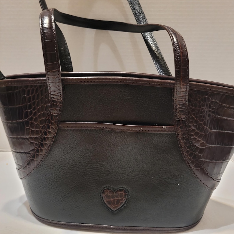 K&ouml;nnte beinhalten: Eine braune und schwarze Handtasche mit herzf&ouml;rmigem Detail. Die Tasche hat ein strukturiertes Krokodilmuster an den Seiten und Griffen, mit einem glatten, dunkelbraunen K&ouml;rper. Sie hat zwei Griffe und einen Schultergurt.