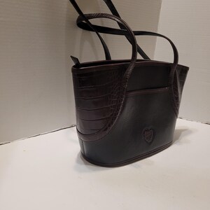 K&ouml;nnte beinhalten: Schwarze und braune Tragetasche mit Herz-Emblem. Die Tasche hat ein strukturiertes Kunstkrokodil-Design auf der Vorder- und Seite, mit einem glatten schwarzen K&ouml;rper. Sie hat zwei Griffe und eine kleine Tasche auf der R&uuml;ckseite.