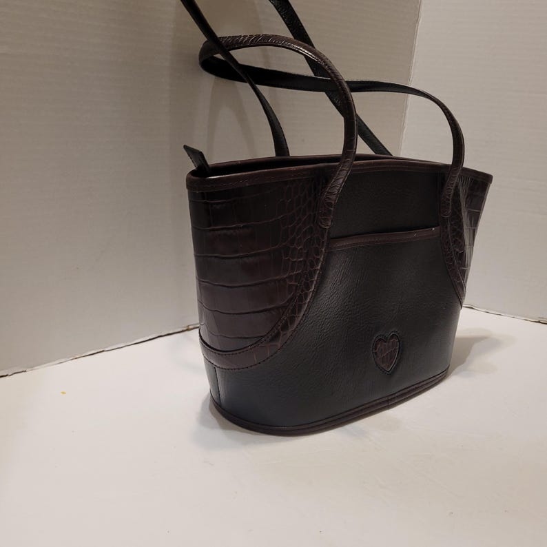 K&ouml;nnte beinhalten: Schwarze und braune Tragetasche mit Herz-Emblem. Die Tasche hat ein strukturiertes Kunstkrokodil-Design auf der Vorder- und Seite, mit einem glatten schwarzen K&ouml;rper. Sie hat zwei Griffe und eine kleine Tasche auf der R&uuml;ckseite.