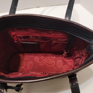 K&ouml;nnte beinhalten: Schwarze und braune Tragetasche mit rotem Innenraum. Die Tasche hat einen Rei&szlig;verschluss und mehrere Innentaschen. Das Innenfutter weist ein dekoratives Wirbelmuster auf. Die Tasche hat zwei schwarze Griffe.