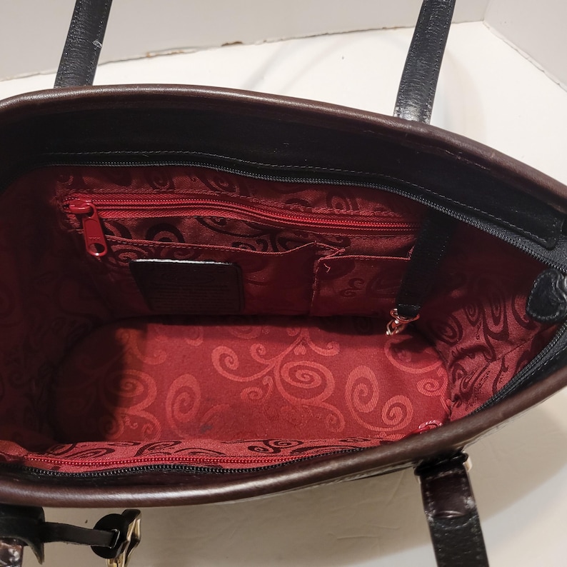 K&ouml;nnte beinhalten: Schwarze und braune Tragetasche mit rotem Innenraum. Die Tasche hat einen Rei&szlig;verschluss und mehrere Innentaschen. Das Innenfutter weist ein dekoratives Wirbelmuster auf. Die Tasche hat zwei schwarze Griffe.