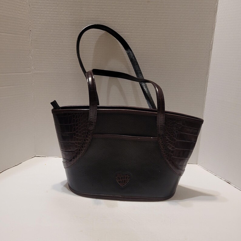 K&ouml;nnte beinhalten: Dunkelbraune Tragetasche mit krokodiltexturierten Akzenten an den Seiten. Die Tasche hat eine glatte, gl&auml;nzende Oberfl&auml;che und ein kleines herzf&ouml;rmiges Emblem auf der Vorderseite. Sie hat zwei Griffe und einen oberen Rei&szlig;verschluss.