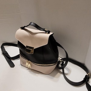 May include: A black and beige backpack with a gold zipper and hardware. The backpack has a top handle and adjustable straps. The brand name "STEVE MADDEN" is visible on the front of the bag.