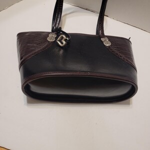 K&ouml;nnte beinhalten: Schwarze und braune Ledertasche mit herzf&ouml;rmigem Anh&auml;nger. Die Tasche hat einen strukturierten braunen oberen Bereich und einen glatten schwarzen K&ouml;rper. Silberfarbene Beschl&auml;ge akzentuieren die Griffe.