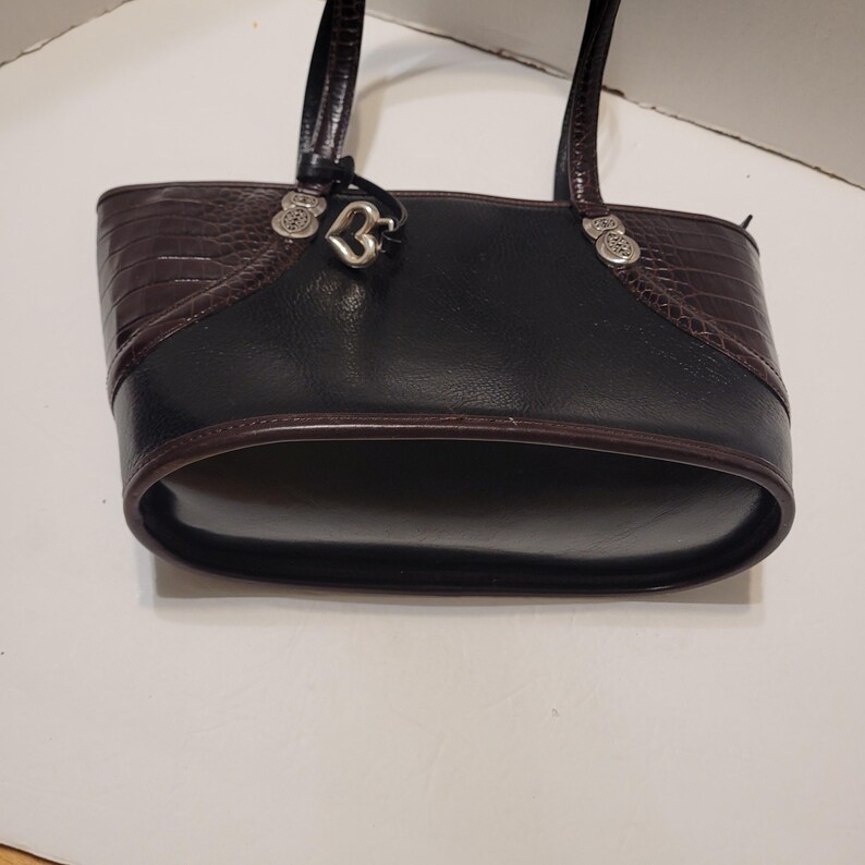 K&ouml;nnte beinhalten: Schwarze und braune Ledertasche mit herzf&ouml;rmigem Anh&auml;nger. Die Tasche hat einen strukturierten braunen oberen Bereich und einen glatten schwarzen K&ouml;rper. Silberfarbene Beschl&auml;ge akzentuieren die Griffe.