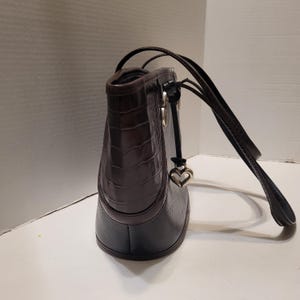 K&ouml;nnte beinhalten: Dunkelbraune Lederhandtasche mit krokodilartiger Textur im oberen Bereich. Die Tasche hat einen schwarzen Boden und einen herzf&ouml;rmigen Anh&auml;nger, der an dem Riemen h&auml;ngt. Die Tr&auml;ger sind dunkelbraun.