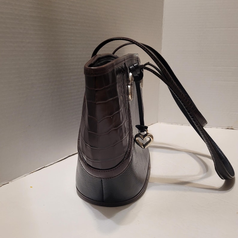 K&ouml;nnte beinhalten: Dunkelbraune Lederhandtasche mit krokodilartiger Textur im oberen Bereich. Die Tasche hat einen schwarzen Boden und einen herzf&ouml;rmigen Anh&auml;nger, der an dem Riemen h&auml;ngt. Die Tr&auml;ger sind dunkelbraun.