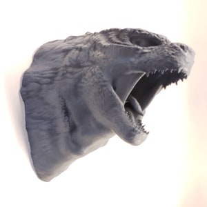 May include: A grey, three-dimensional wall-mounted sculpture of a creature head with a textured, scaly surface. The creature has an open mouth revealing sharp, pointed teeth and a visible tongue, with a rugged, uneven skin texture throughout.