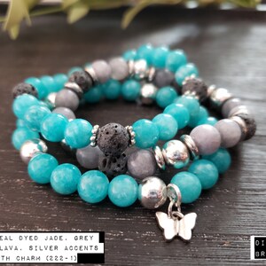 Op de afbeelding: Drie-strengs diffuser armband met teal geverfde jade, grijze jade, lava kralen en zilveren accenten. De armband heeft een vlinderbedel en de tekst "#1 8MM TEAL DYED JADE, GREY JADE, LAVA, SILVER ACCENTS WITH CHARM (222-1) DIFFUSER BRACELET" is zichtbaar.
