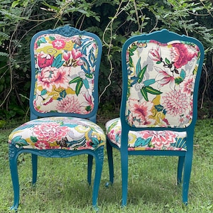 Pode incluir: Duas cadeiras azul-petr&oacute;leo com estofamento estampado floral. As cadeiras t&ecirc;m detalhes talhados ornamentados nas costas e nas pernas. As cadeiras est&atilde;o sentadas em um peda&ccedil;o de grama verde.