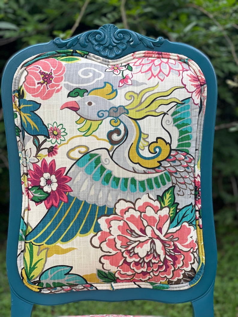 Pode incluir: Cadeira azul turquesa com um assento de tecido estampado com flores que apresenta um design de p&aacute;ssaro colorido. A cadeira tem uma parte superior de madeira esculpida e um encosto curvo.