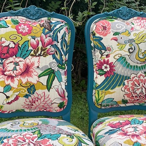 Pode incluir: Duas cadeiras azul-petr&oacute;leo com encostos talhados ornamentados e estofamento com padr&atilde;o floral. As cadeiras t&ecirc;m um fundo branco com desenhos florais rosa, amarelo, verde e azul.