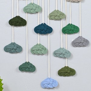 Könnte beinhalten: Ein Wandbehang mit einer Holzleiste und weißem Garn mit 15 geknüpften Wolken in Grün-, Blau- und Grautönen.
