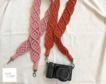 May include: Two macrame camera straps in dusty pink and burnt orange, featuring a repeating diamond knot pattern. The orange strap is attached to a black Sony camera with metal swivel clips, while the pink strap lies beside it with matching clips.