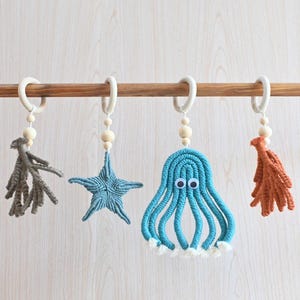 May include: Four macrame hanging ornaments suspended from a wooden rod. The set includes a grey coral shape, a blue starfish, a blue octopus with googly eyes and white fringe, and an orange coral shape, each attached to a wooden ring and beads.