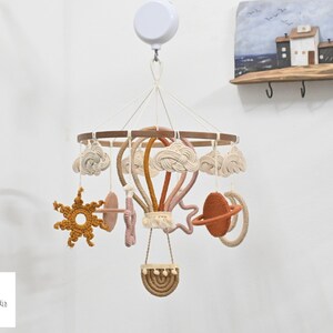 May include: A wooden baby mobile with a hot air balloon, clouds, a sun, a star, and a planet. The mobile is made of macrame and has a neutral color palette of beige, brown, and pink.