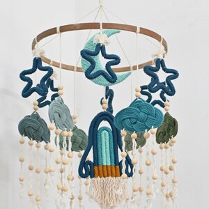 May include: A blue and green baby mobile with a rocket, clouds, stars, and a crescent moon. The mobile is made of yarn and has wooden beads hanging from it.