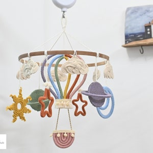 Może przedstawiać: Kolorowy mobil dla dziecka z balonem na gorące powietrze w tęczy, chmurami, gwiazdami i planetą. Mobil jest wykonany z makramu i ma drewniany pierścień.
