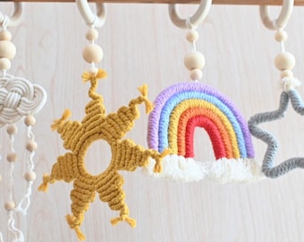 May include: Four macrame hanging ornaments suspended from a wooden rod. The set includes a cream cloud with beaded fringe, a mustard yellow sun, a multi-coloured rainbow with white clouds, and a grey star, each attached to a wooden ring.