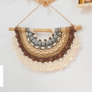 May include: A brown, beige and grey macrame wall hanging with a wooden dowel at the top. The hanging is in the shape of a half-circle and has a fringe at the bottom.