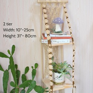 May include: A two-tier macrame hanging shelf with wooden shelves. The shelf is decorated with floral accents and holds a small potted plant, books, and a decorative mushroom. Width is 10 inches to 25 cm, height is 31 inches to 80 cm.
