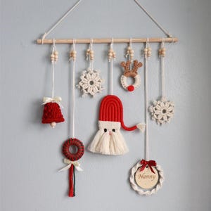 May include: A handmade Christmas wall hanging featuring a wooden dowel with hanging ornaments. The ornaments include a red bell, snowflakes, a reindeer, a Santa Claus, and a wreath. The word "Nanny" is on a wooden circle.