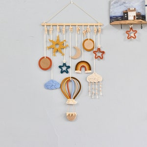 Könnte beinhalten: Ein Makramee-Wandbehang mit einem Regenbogen, einem Heißluftballon, Wolken, Sternen und einer Sonne. Der Wandbehang ist aus Naturfasern gefertigt und hat einen rustikalen, bohemischen Stil.